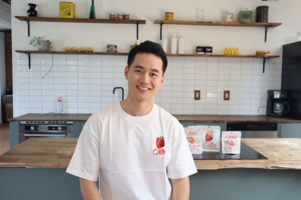 Han Yue, founder of OHME! Foods Inc. wears a white t-shirt with the company logo, posing in front of a pantry. OHME! products are placed on top of the counter.